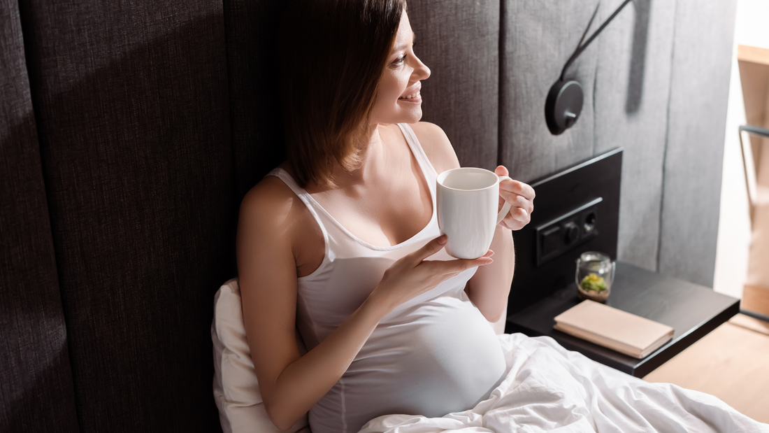 Pregnant woman drinking a warm beverage in bed, smiling.